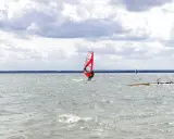 Czerwony żagiel do windsurfingu na wodzie, surfer w akcji. W tle inni windsurferzy i chmury.