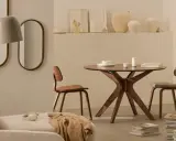 Dos espejos ovalados cuelgan sobre un sofá beige. A la derecha, una mesa redonda de madera con sillas y un jarrón.