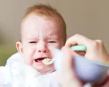 Płacz rocznego dziecka, które nie chce jeść samodzielnie. Mama próbuje nakarmić je łyżeczką.
