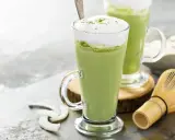 Dwa zielone napoje matcha w szklankach z pianką, obok bambusowa miotełka i wiórki kokosowe. Tak smakuje matcha.