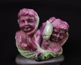 Due putti scolpiti in pietra, uno con la mano sul mento, sembrano abbracciare un elemento verde, forse un fiore. La pietra, un rubino verde, dona un tocco di magia.