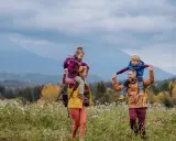 Rodzina na spacerze po łące. Dzieci siedzą na ramionach rodziców, jakby hasające zające zakopane w trawie.