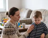 Mama uspokaja dziecko, które wpada w histerię z byle powodu. W tle widać kanapę i stolik z zabawkami.