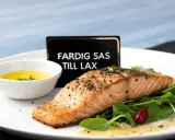En perfekt tillagad laxfilé serveras med en skål med gul sås till lax, gröna blad och en röd bär.