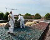 Dwóch pracowników w kombinezonach ochronnych usuwa z dachu stare płyty eternitowe.