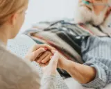 Mani che si stringono tra una giovane donna e un anziano a letto, con cannule nasali. Il caregiver viene pagato per la sua dedizione.