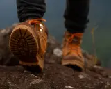 Stopy w brązowych butach trekkingowych z pomarańczowymi sznurówkami na kamienistym szlaku. Membrana w butach zapewnia komfort podczas wędrówki.