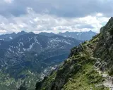 Kamienista ścieżka wije się wzdłuż skalistego grzbietu w Tatrach Wysokich. W oddali widać ośnieżone szczyty i jezioro.