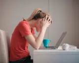 Kobieta z rękami na głowie przy laptopie, kubek i słuchawki. Czy to objawy wypalenia zawodowego jak sobie radzić?