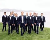 Groomsmen in navy suits with bow ties walk on grass by a lake. Can you wear a normal suit to a black tie event? This image shows a stylish alternative.