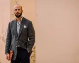 Man in a textured grey jacket and black pants, holding a tan envelope clutch.