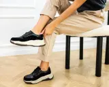 Kobieta w beżowych spodniach i czarnych sneakersach na grubych podeszwach. Pokazuje, jak szybko wysuszyć buty, by znów cieszyć się komfortem.