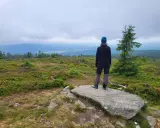 Osoba na skale podziwia rozległy widok z wysokiej kopy. W oddali widać pasma górskie i lasy.