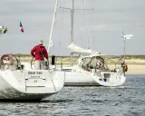 Un homme en gilet rouge tient la barre d'un voilier, le pont est bien mouillé. Un autre voilier suit, naviguant sur une mer calme.