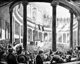 Versammlung der Frankfurter Nationalversammlung in der Paulskirche. Viele Männer in Anzügen und Hemden sitzen und stehen in einem großen, säulenreichen Gebäude.