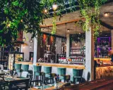Wnętrze hotelu z browarem: bar z zielonymi hokerami, miedziany destylator, półki z butelkami i mnóstwo zieleni.