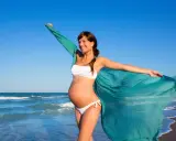 Donna incinta in bikini bianco sulla spiaggia, con un velo turchese che svolazza al vento. Il mare sullo sfondo.
