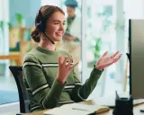 Uśmiechnięta customer experience manager w słuchawkach gestykuluje, siedząc przy biurku przed monitorem.