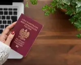 Dłoń trzyma polski paszport na tle laptopa i roślin.