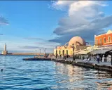Il porto di Chania con il suo faro iconico e la moschea ottomana, bagnato da un mare blu intenso sotto un cielo nuvoloso.