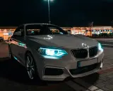 Białe BMW z włączonymi światłami adaptacyjnymi błyszczy nocą przed sklepami.