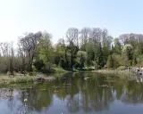 Spokojny staw w Bolestraszycach, otoczony drzewami i zielenią. Widoczna drewniana kładka z ludźmi, idealne miejsce na relaks i podziwianie atrakcji.