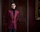 A man in a patterned burgundy jacket and black turtleneck, perfect for cocktail party attire for men, stands in a dimly lit room with ornate wooden doors.