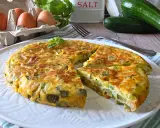 Una gustosa frittata di zucchine, dorata e soffice, servita su un piatto bianco. Ingredienti freschi come uova e zucchine sono visibili sullo sfondo.