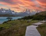 Drewniana ścieżka prowadzi przez zielone wzgórza ku turkusowemu jezioru i ośnieżonym górom. Zachód słońca maluje niebo nad **Tierra del Fuego** odcieniami różu i pomarańczy.