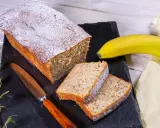 Ein **super saftiger Bananenkuchen**, bestäubt mit Puderzucker, liegt auf einem schwarzen Stein neben zwei Scheiben. Eine Banane und ein Messer liegen daneben.