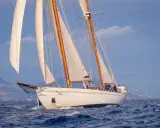 Un élégant voilier blanc, tel un cygne noir sur l'eau, navigue avec ses voiles gonflées par le vent, longeant une côte méditerranéenne.