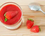 Un verre de panna cotta avec une fraise entière sur un coulis de fraise. Deux fraises fraîches et une cuillère à côté.