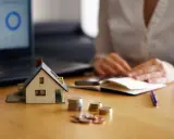Maqueta de casa, monedas y portátil con gráfico. Persona anotando en libreta. Analizando el precio vivienda.