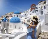 Una madre abbraccia la sua bambina, ammirando il panorama di Santorini, Grecia. Le cupole blu brillano sotto il sole, mentre le case bianche si aggrappano alla collina.