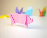 Cerdo de origami rosa con otras figuras de papiroflexia desenfocadas al fondo.