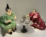 Dos figuras voluminosas, al estilo de Niki de Saint Phalle, sentadas a una mesa de té. Una viste de verde, la otra de rojo.