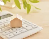 Maison en bois sur une calculatrice blanche, symbolisant le calcul du prix du **pel plafond**. Des feuilles vertes floues en arrière-plan.