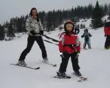 Kobieta na nartach trzyma szelki, za którymi jedzie dziecko. W tle inni narciarze.