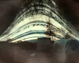 Paesaggio distorto con alberi e strutture, catturato con un **foro stenopeico**. Le strisce luminose suggeriscono un lungo tempo di esposizione.