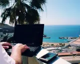 Praca na Cyprze: człowiek pisze na laptopie z widokiem na morze i palmę. Idealne miejsce na zdalną pracę.