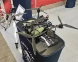 Ich sehe einen großen, schwarzen Quadrocopter mit grünen Zylindern, der auf einem Tisch steht. Die Propeller sind aus Carbon.