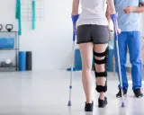 Fizjoterapeuta uczy, jak chodzić o kulach, wspierając pacjentkę z ortezą na nodze w sali rehabilitacyjnej.