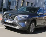 Ciemnoszary Toyota RAV4 Hybrid pędzi po mieście, odbijając w lustrach budynki i niebo.