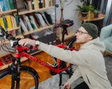Mężczyzna przygotowuje rower do wysyłki w całości. W tle regał z książkami i rośliny.