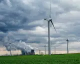 Windräder auf grüner Wiese unter dramatischem Himmel, im Hintergrund rauchende Industrieanlagen. Ein Symbol für Investitionen in erneuerbare Energien in Deutschland.