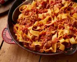 En stor portion pasta bolognese med breda bandnudlar och en mustig köttfärssås i en röd gjutjärnspanna.