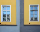 Dwa drewniane okna na żółtej ścianie, jedno odnowione, drugie czekające na renowację.
