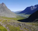 Vacker vy över Sareks nationalpark med Rapadalen i fjärran. En flod slingrar sig genom en grön dal omgiven av höga, steniga berg under en klarblå himmel.