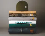 Stos ubrań Carhartt: czapka, bluzy i jeansy. Carhartt to firma znana z wytrzymałej odzieży roboczej i streetwearowej.