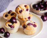 Saftige Muffins mit Sauerkirschen, bestäubt mit Puderzucker. Frische Kirschen liegen daneben und auf einem Muffin.